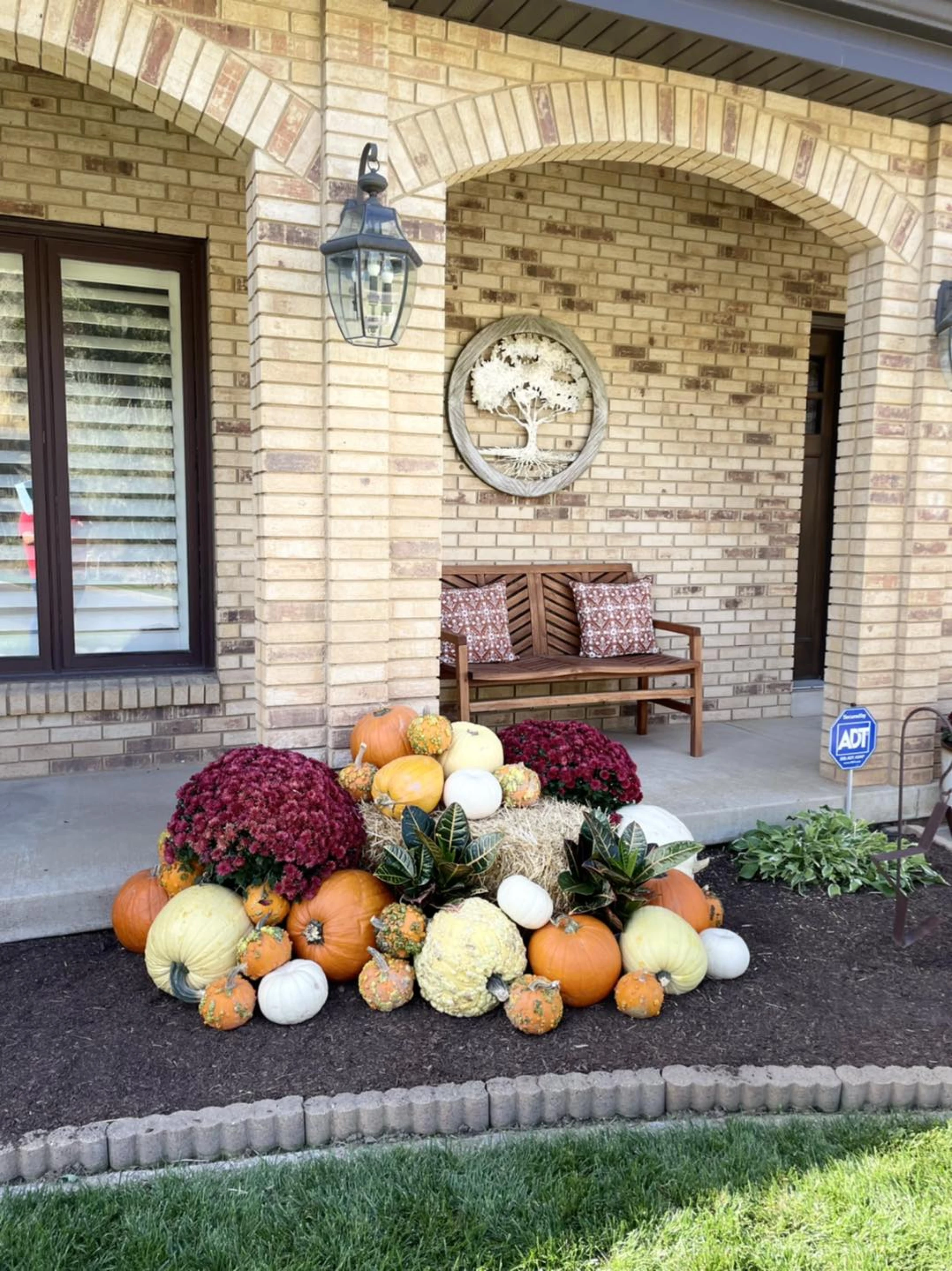 Fall porch display with pumpkins, hay bales, and mums in front of a brick house with a wooden bench